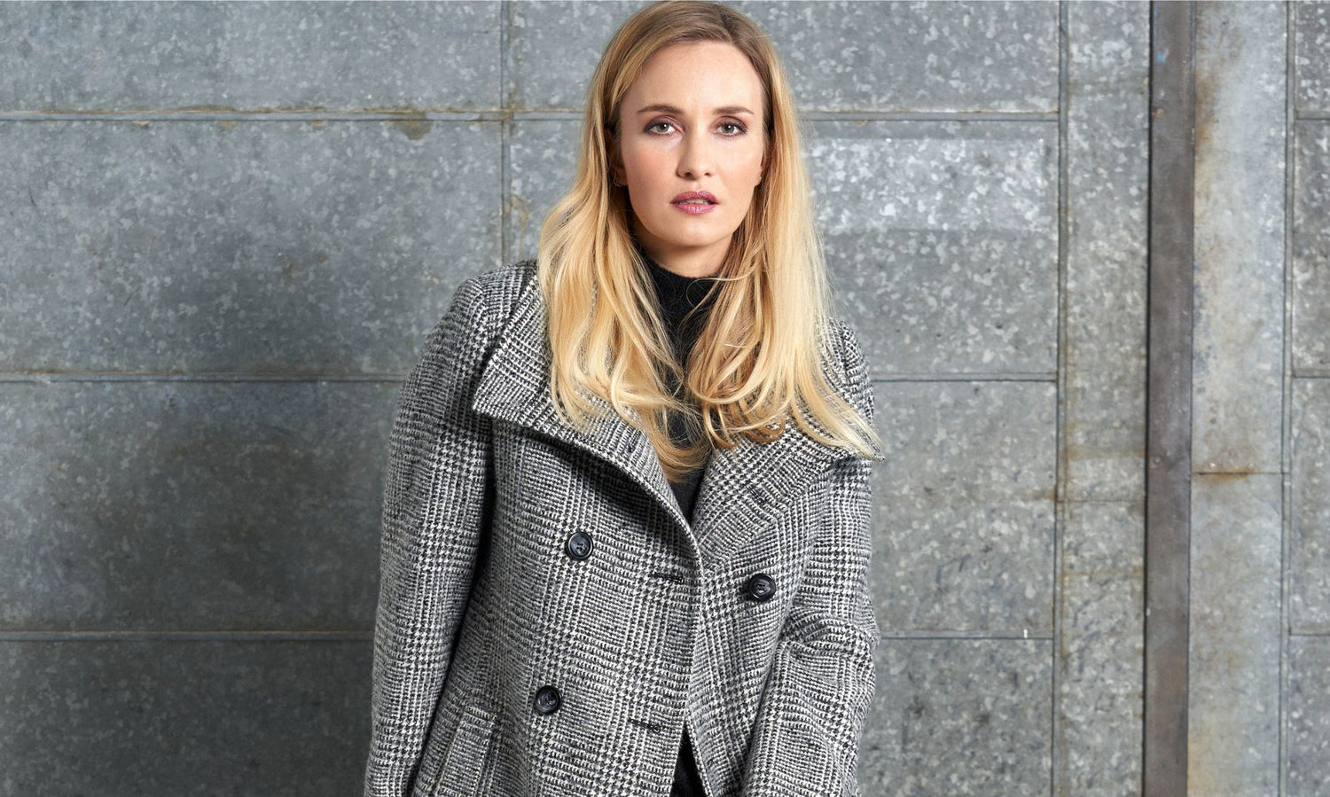 Kvinna iförd en gråmelerad ulljacka från Junge står framför en metallisk bakgrund. Exempel på ulljackor och ullkappor för damer från Junge